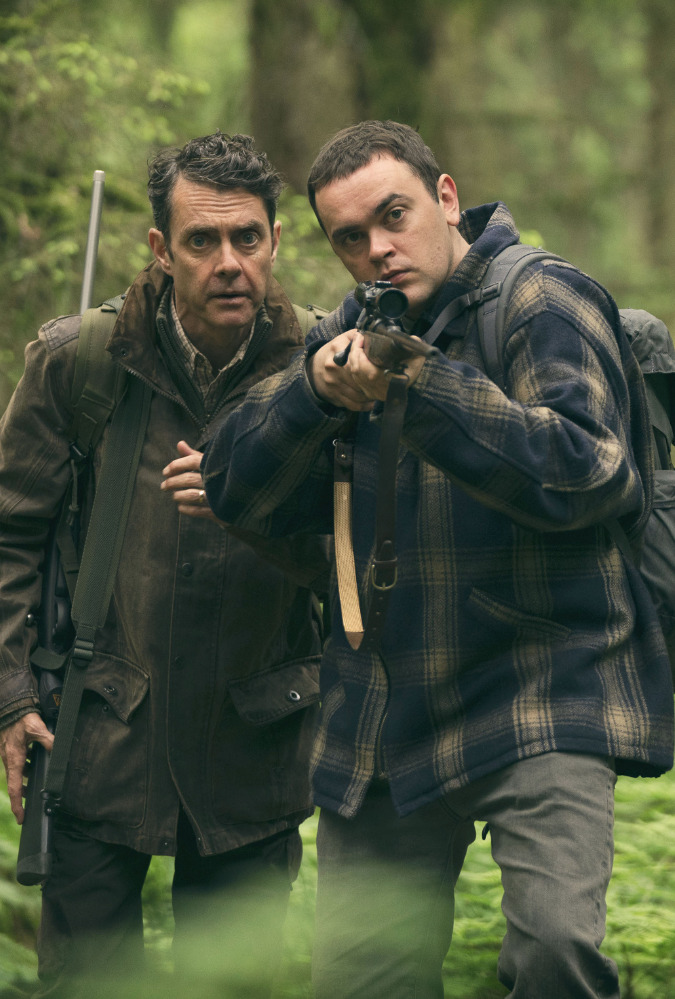 Still from Kill. Man and his son in his twenties stand in a wood. The son holds a rifle out in front of him which they both stare through.
