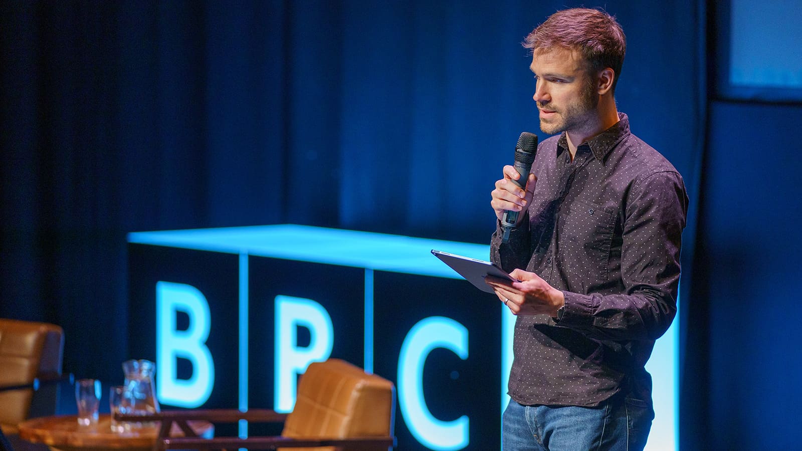 A man stands on a stage speaking into a microphone. Behind him is a large version of the BBC logo.