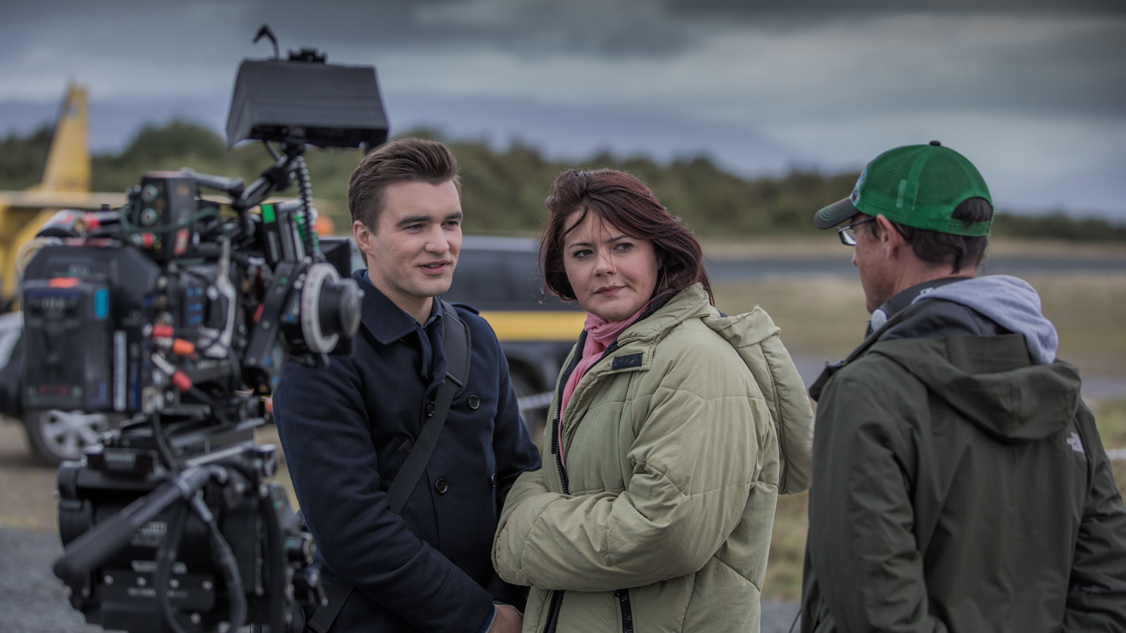 Two actors on a beach filming Bannan on location - there is a camera in front of them and they are talking to a member of the crew