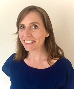 Katharine Simpson stands smiling at the camera - we can see her head and shoulders as she stands wearing a navy top against a light cream wall. She has longer light brown hair.