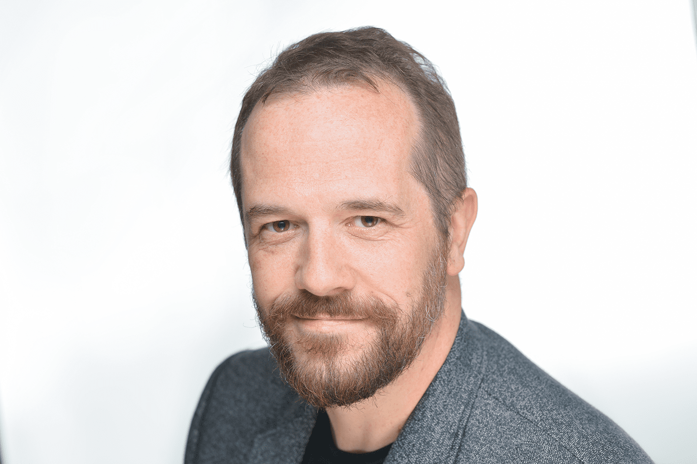 Mark is a man with short hair and a short beard - he sits in front of a white background, wearing a light greyish blue top