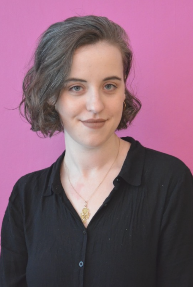 Anna stands wearing a black top against a bright pink background. She has short hair and gently smiles at the camera.