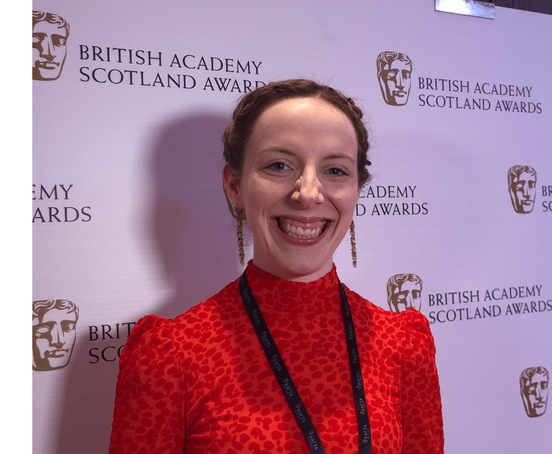 Eilidh wears a red dress and smiles - she's standing against a backdrop at an event with various logos