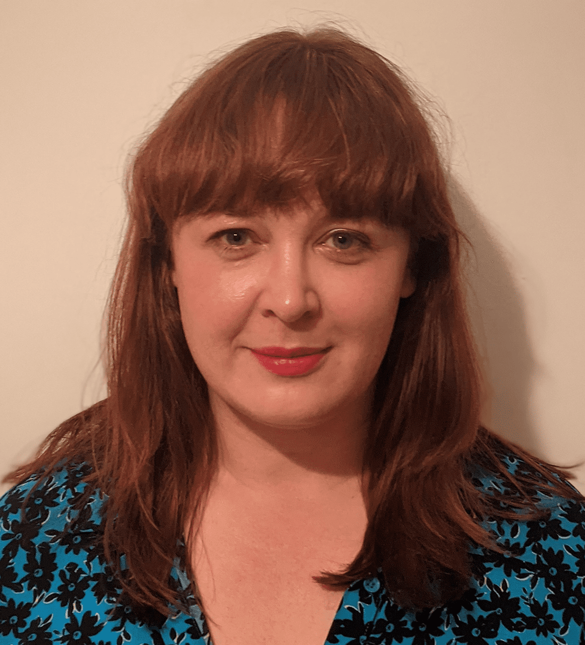 Louise stands against a cream wall - she has dark red hair and wears a blue top