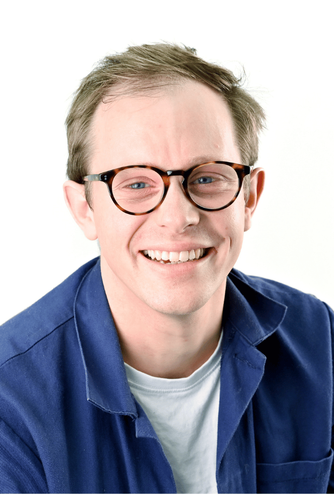 Samuel wears a dark blue shirt over a white t-shirt and wears tortoiseshell framed round glasses