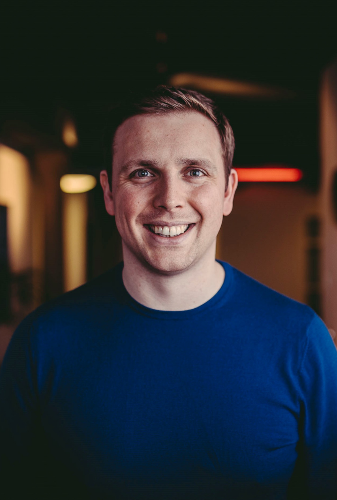 Steven stands in a room with lamps behind him wearing a dark blue jumper