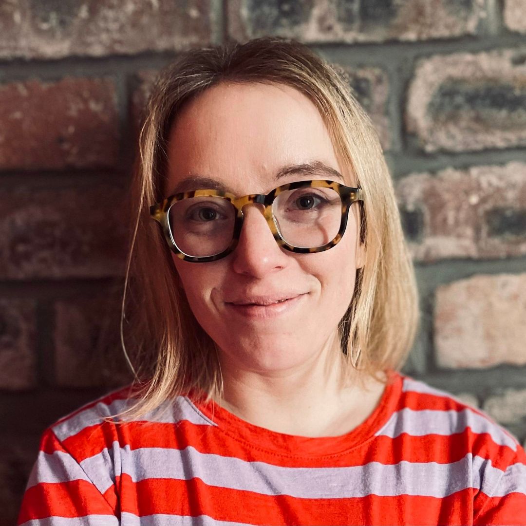 A woman with shoulder length blonde hair and glasses smiles at the camera, She wears a red striped top.