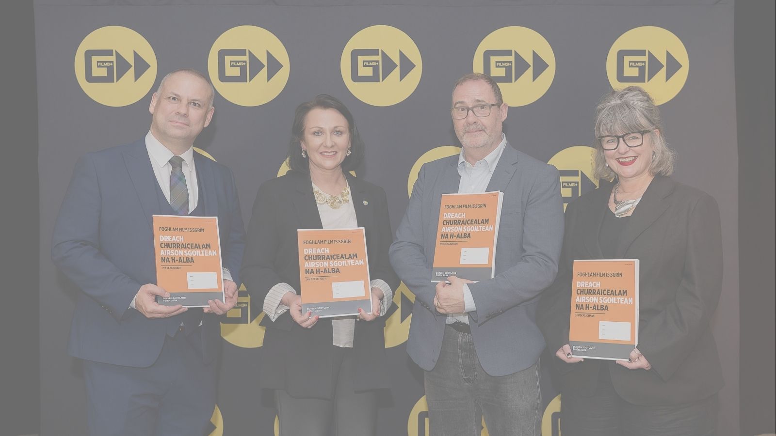 Four people stand in front of a backdrop featuring yellow logos, each holding an orange booklet titled 