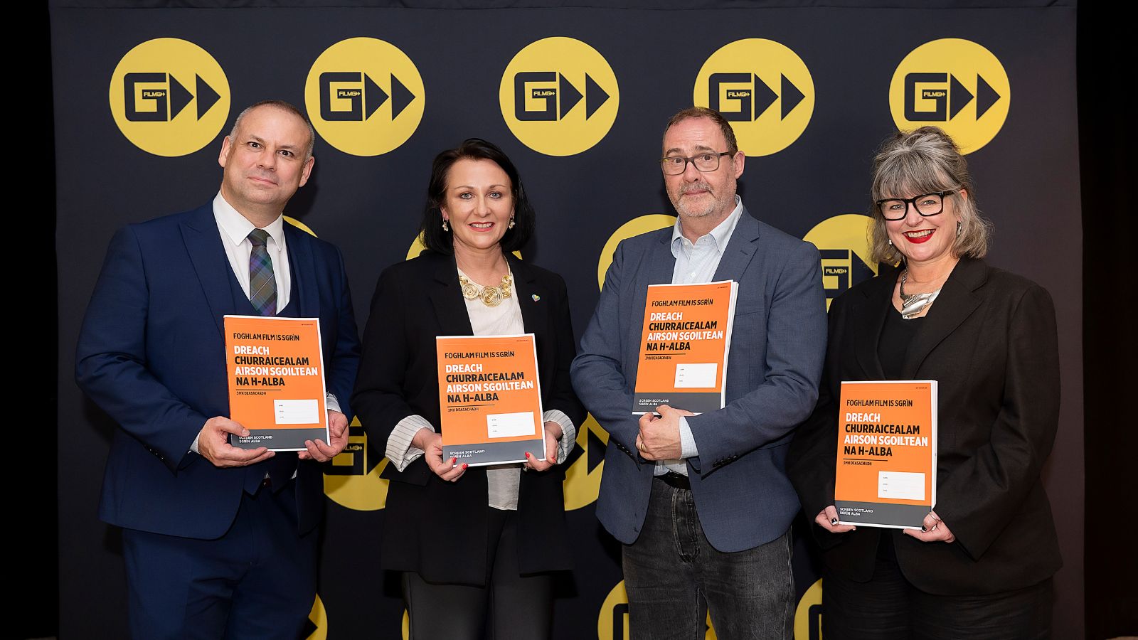 Four people stand in front of a backdrop featuring yellow logos, each holding an orange booklet titled 