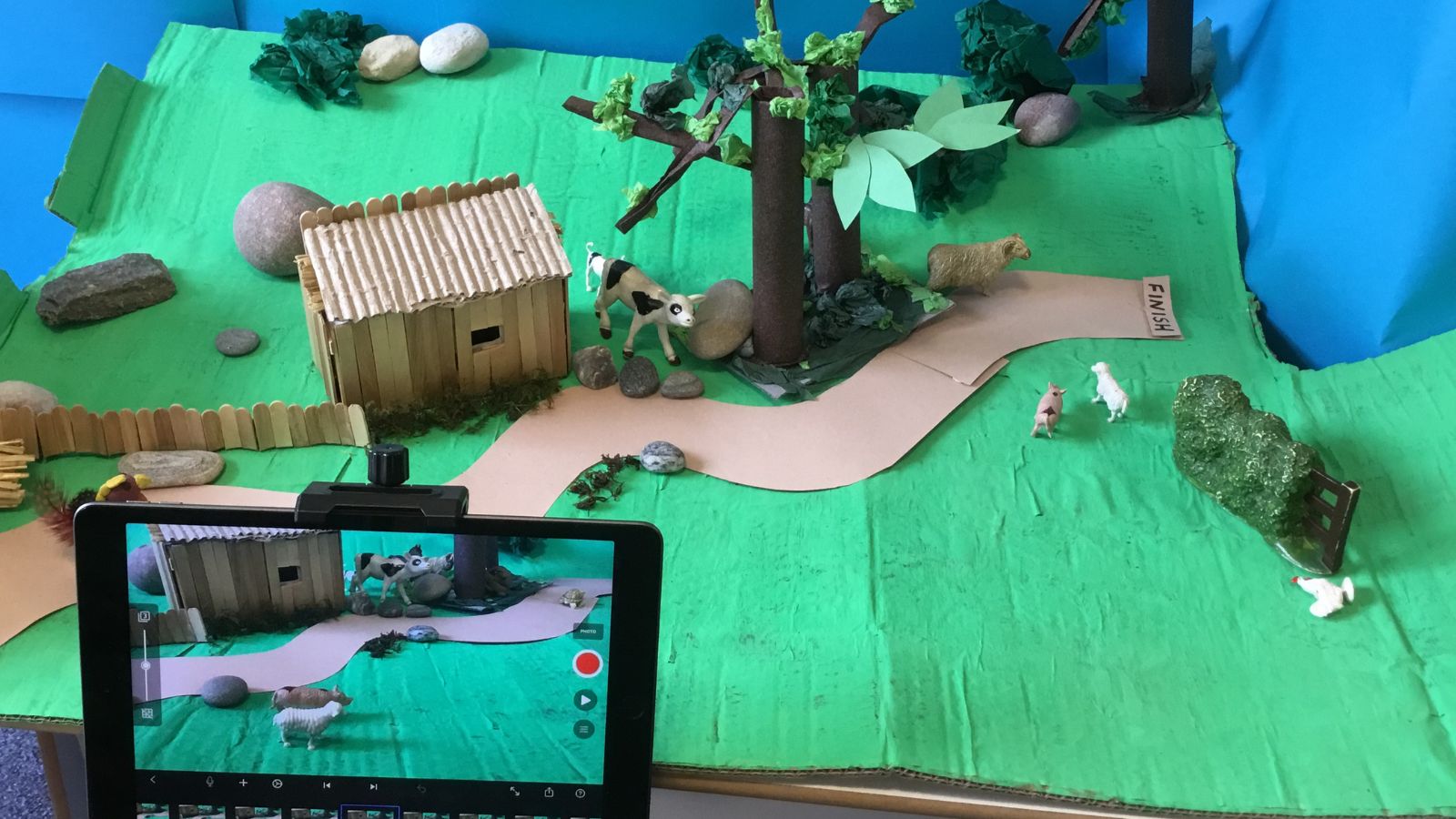 A diorama on a green base depicts a rural scene with a small wooden structure resembling a barn. There are miniature figures of a cow, goat, and pig on a paper path leading to a "FINISH" sign. A tablet in the foreground displays a live view of the scene, suggesting video recording. Trees and rocks create a naturalistic setting.