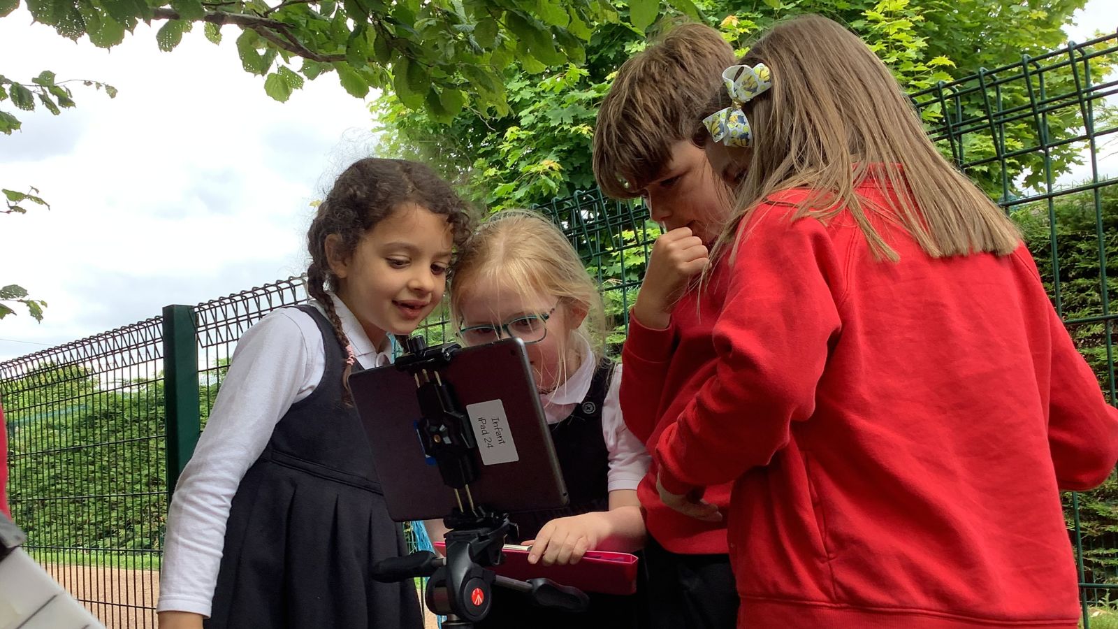 Four children are gathered around an iPad mounted on a tripod outdoors. They are engaged and looking intently at the screen, surrounded by a fence and lush green leaves.