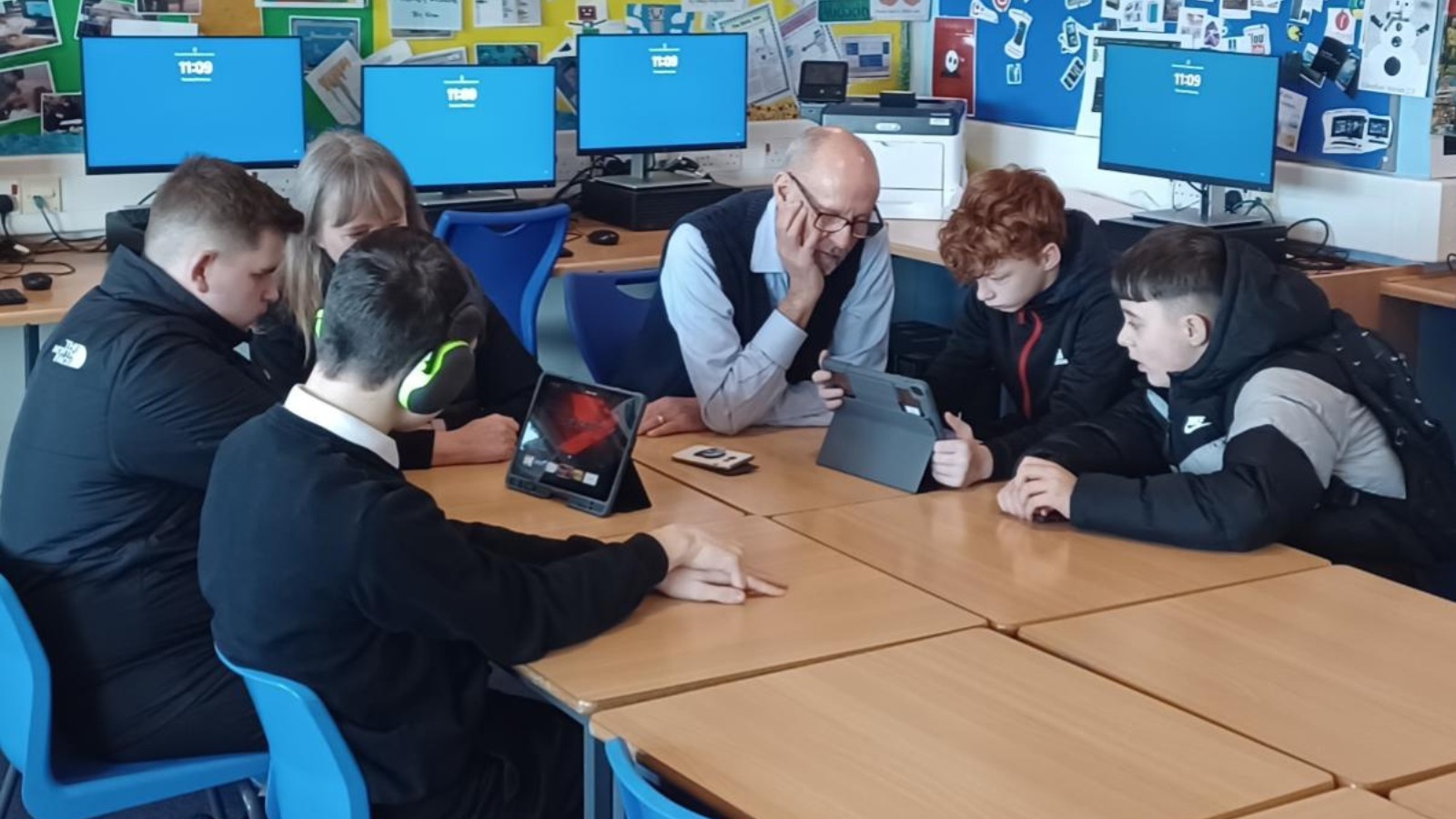 School pupils sit around a desk, looking at iPads, with a teacher in a classroom