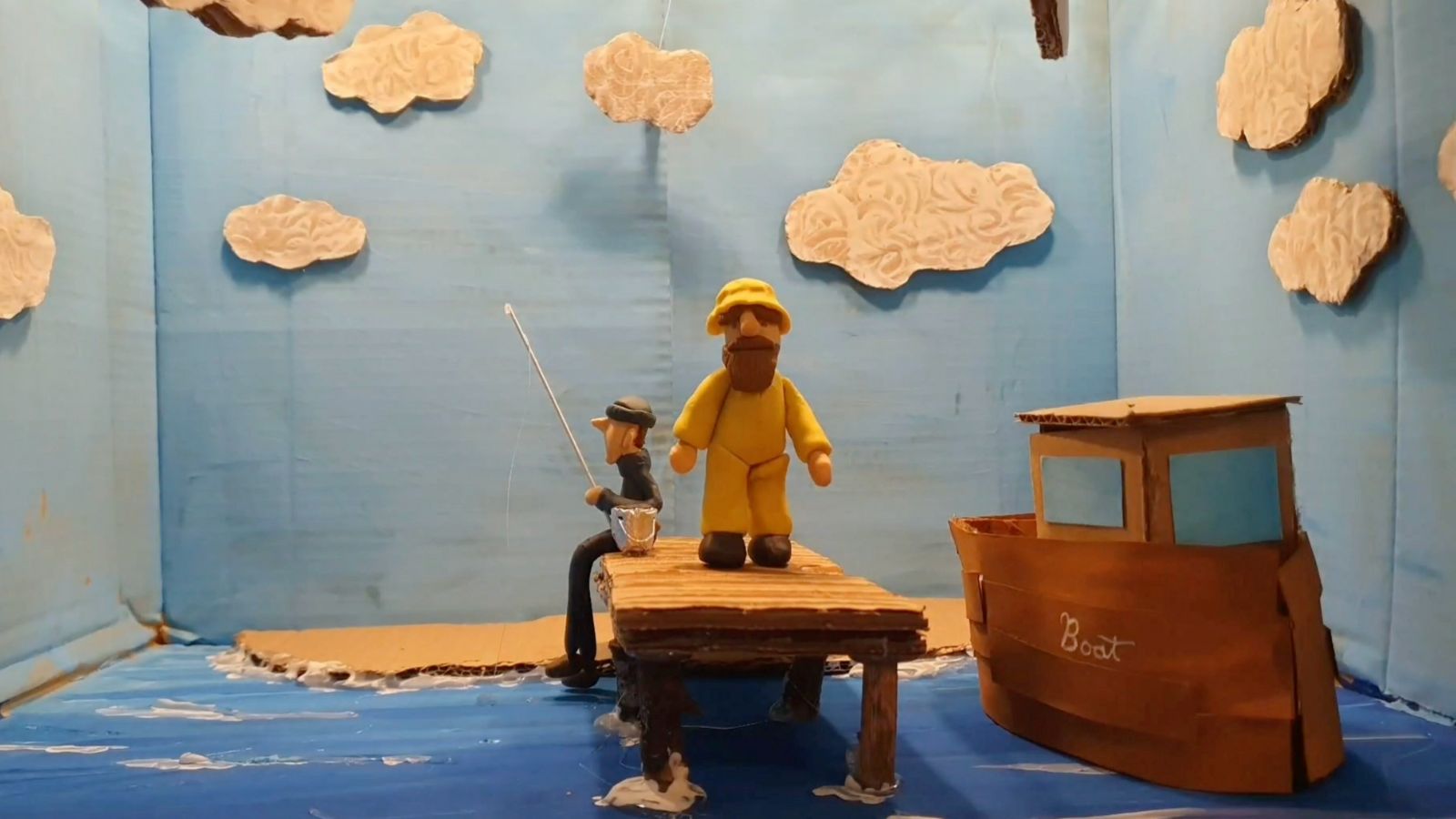 Clay figures of two men are on a dock in a blue diorama. One man wears yellow rain gear, and the other, in black, is fishing. A brown cardboard boat labeled 