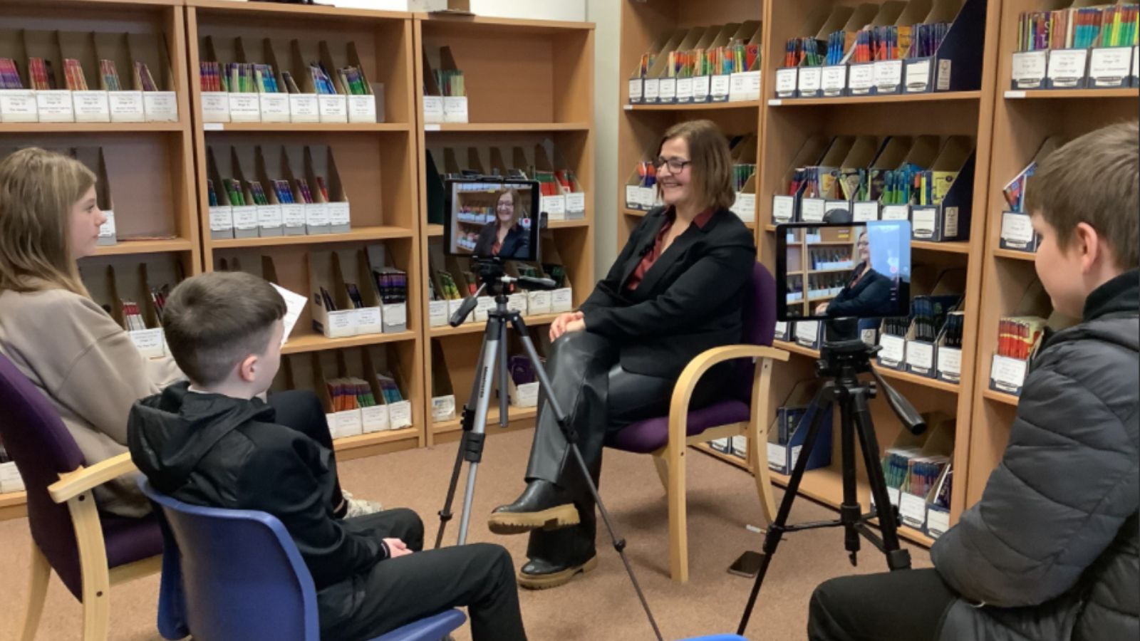 Four people are seated in a library filled with books, having a conversation. Two children sit facing a woman in a black suit. Two tablets on tripods display the same woman. Shelves with colourful book collections are neatly organised in the background.