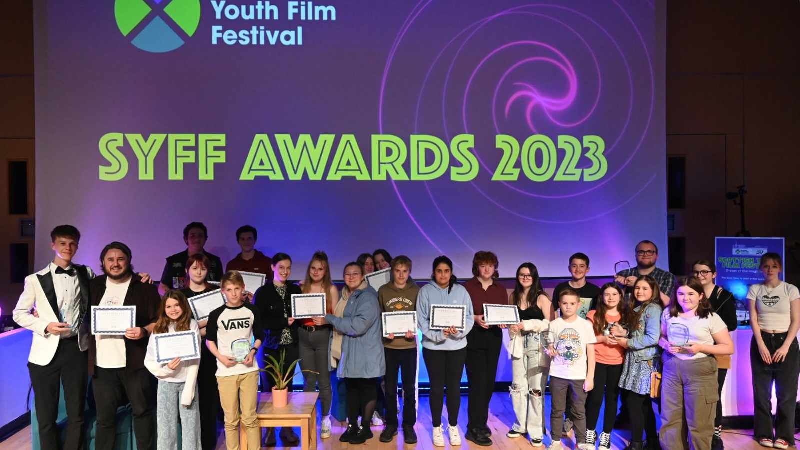 A group of children and teenagers stand in two rows holding certificates. Behind them a screen reads SYFF Awards 2023.