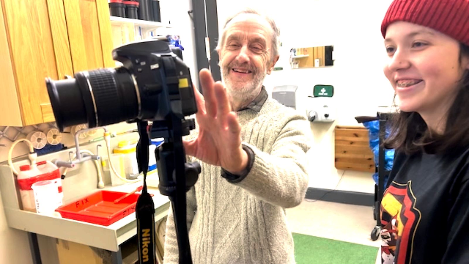 An elderly man with a beard and gray sweater is standing beside a camera on a tripod, smiling while adjusting it. Next to him, a girl with braces and wearing a red beanie and graphic T-shirt smiles broadly. The background includes shelves and a table with photography supplies.