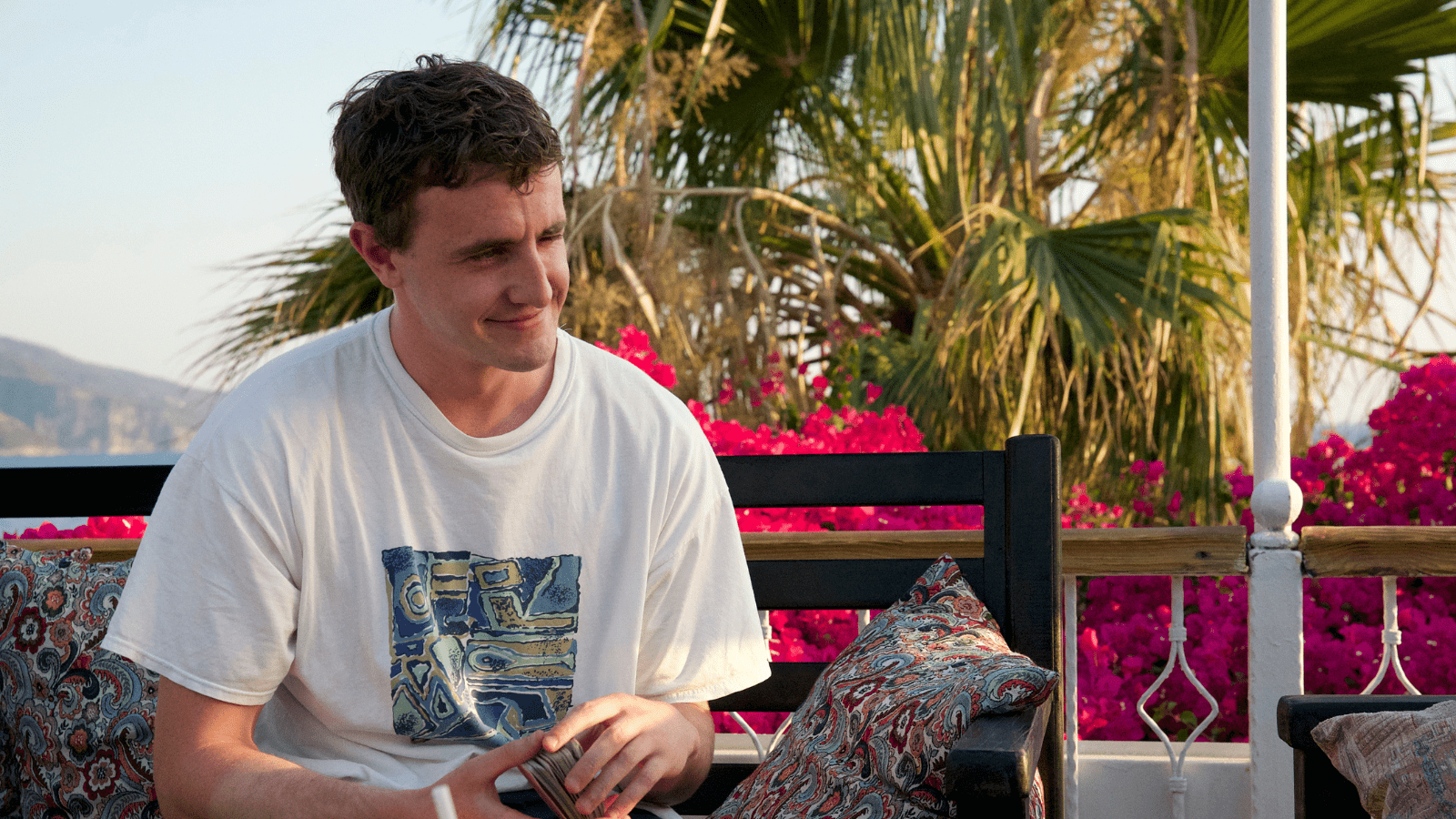 Aftersun still. Paul Mescal character smiles while sitting down outside. Pink blossoms can be seen behind him
