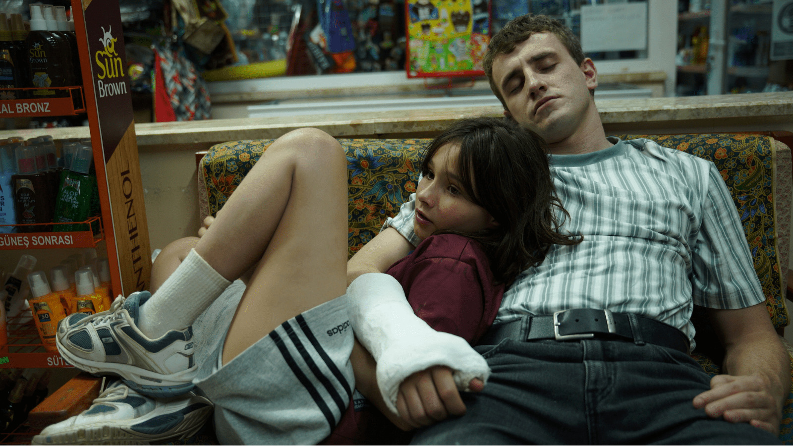 A man with a broken wrist in a cast holds a young girl, his daughter, as they lie on a sofa - this is a still from Aftersun, with Paul Mescal and Frankie Corio playing father and daughter respectively