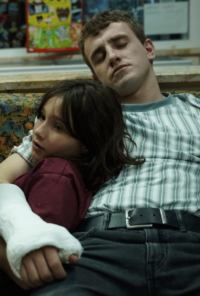 A man with a broken wrist in a cast holds a young girl, his daughter, as they lie on a sofa - this is a still from Aftersun, with Paul Mescal and Frankie Corio playing father and daughter respectively