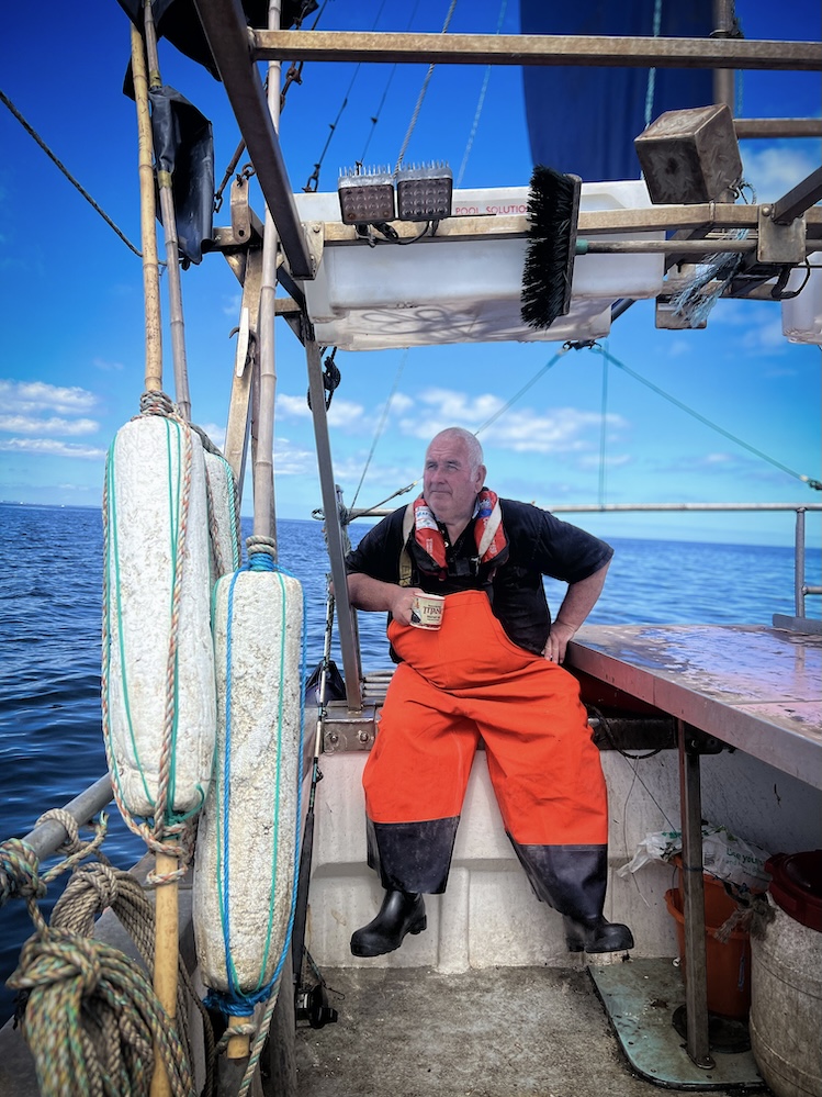 A man in bright orange overalls and a life jacket stands on a fishing boat, holding a mug. He gazes into the distance over the calm blue ocean under a clear sky with scattered clouds. The boat's deck and safety equipment are visible in the foreground.