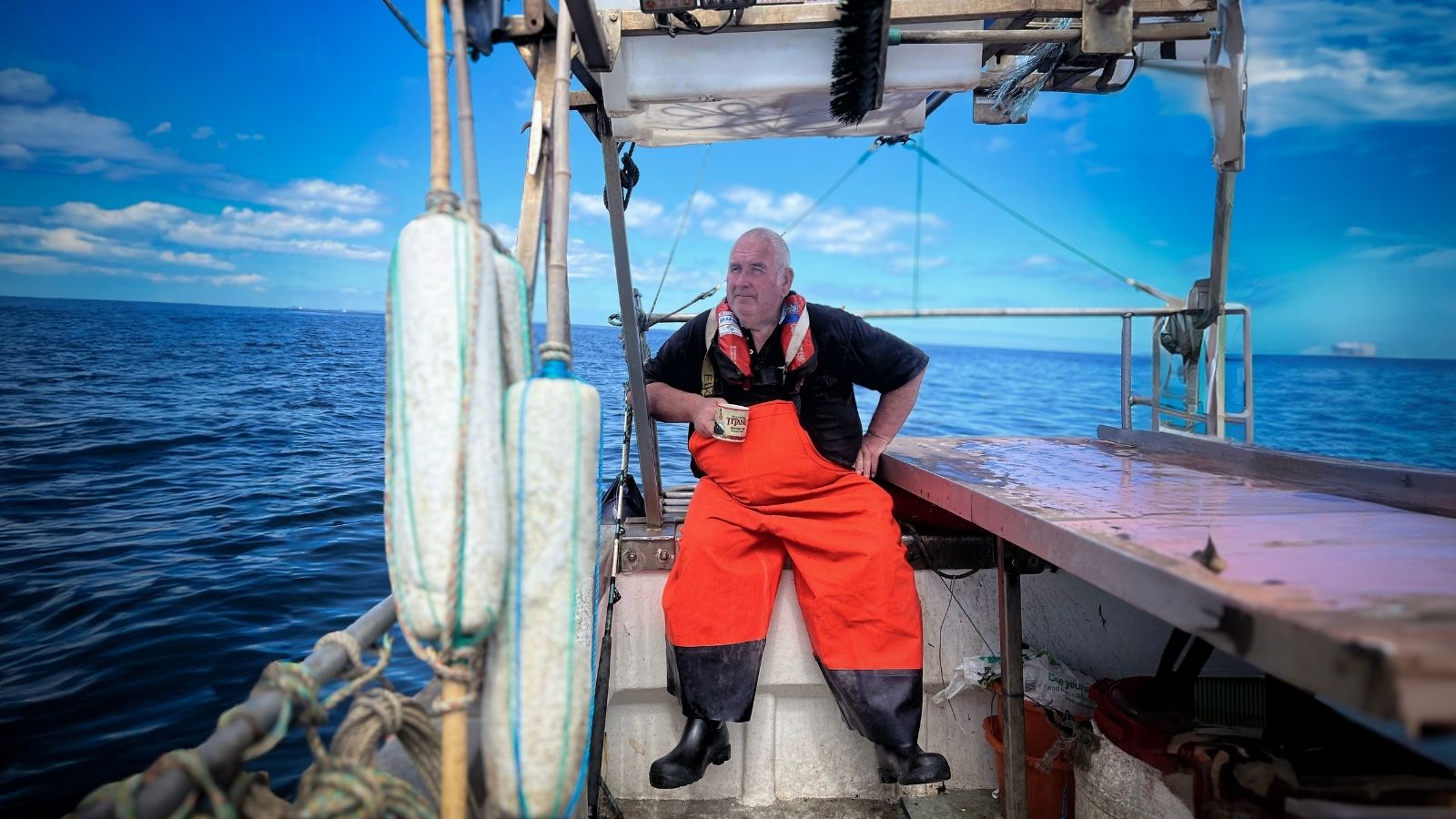 A man in bright orange overalls and a life jacket stands on a fishing boat, holding a mug. He gazes into the distance over the calm blue ocean under a clear sky with scattered clouds. The boat's deck and safety equipment are visible in the foreground.