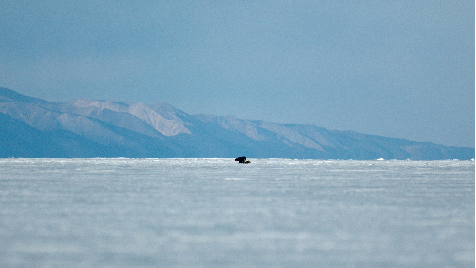 A wide landscape shot of a person in the distance kneeling on snow