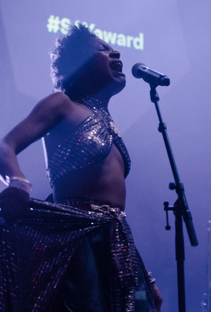 A performer passionately sings into a microphone on a dimly lit stage, wearing a shimmering, metallic outfit. The background shows purple lighting.