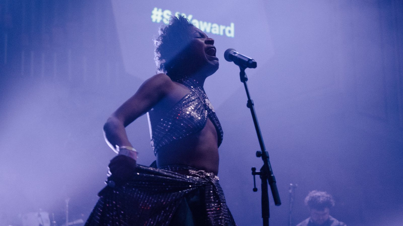 A performer passionately sings into a microphone on a dimly lit stage, wearing a shimmering, metallic outfit. The background shows purple lighting.