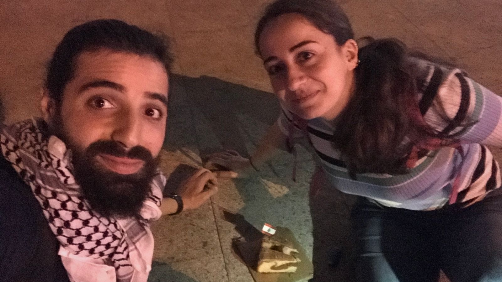 Two people sitting on the ground at night, smiling at the camera. A small piece of cake with a toothpick flag decoration is placed on the ground between them. The lighting is dim and warm, casting shadows.