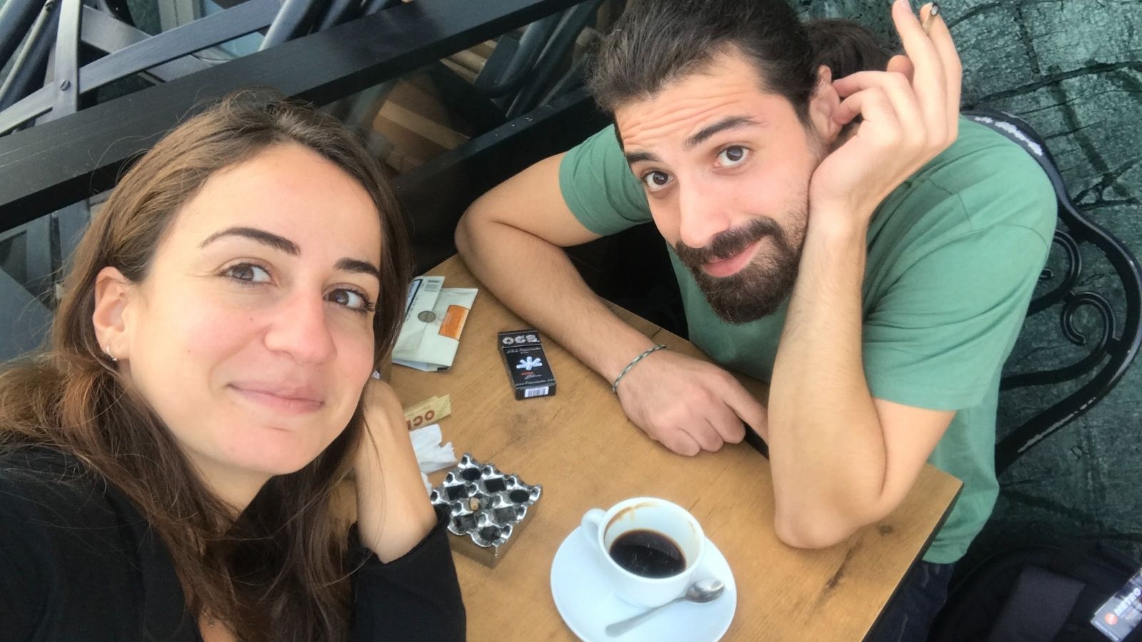 Two people sitting at a wooden table, smiling at the camera. One person is holding a cigarette, and a cup of black coffee sits in front of them along with cigarette packs and a lighter. They are seated outdoors on a metal chair, with a structural backdrop.