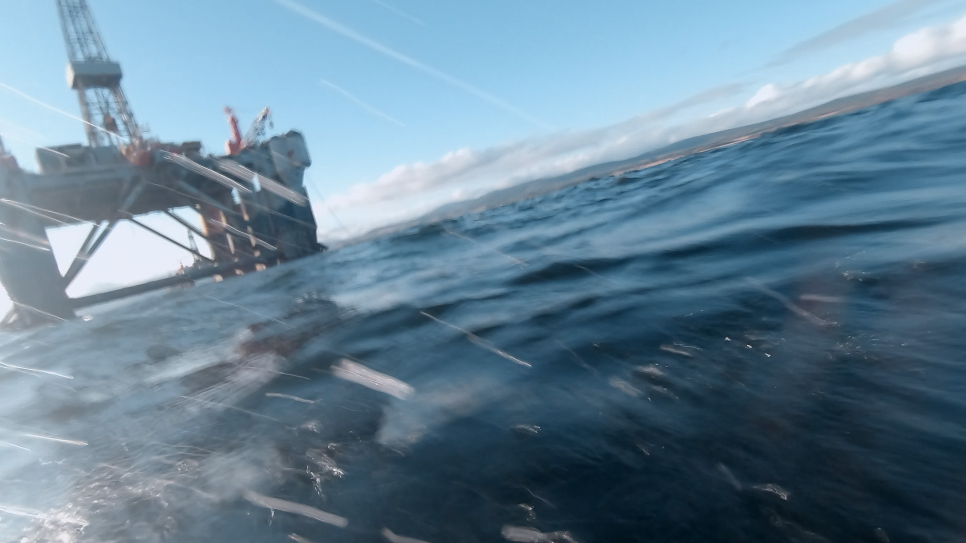 Still from The Oil Machine. An Oil Rig can be seen in the sea. The photo is slightly blurred with water splashing up on the camera lense, appearing as if the image is taken from a boat.