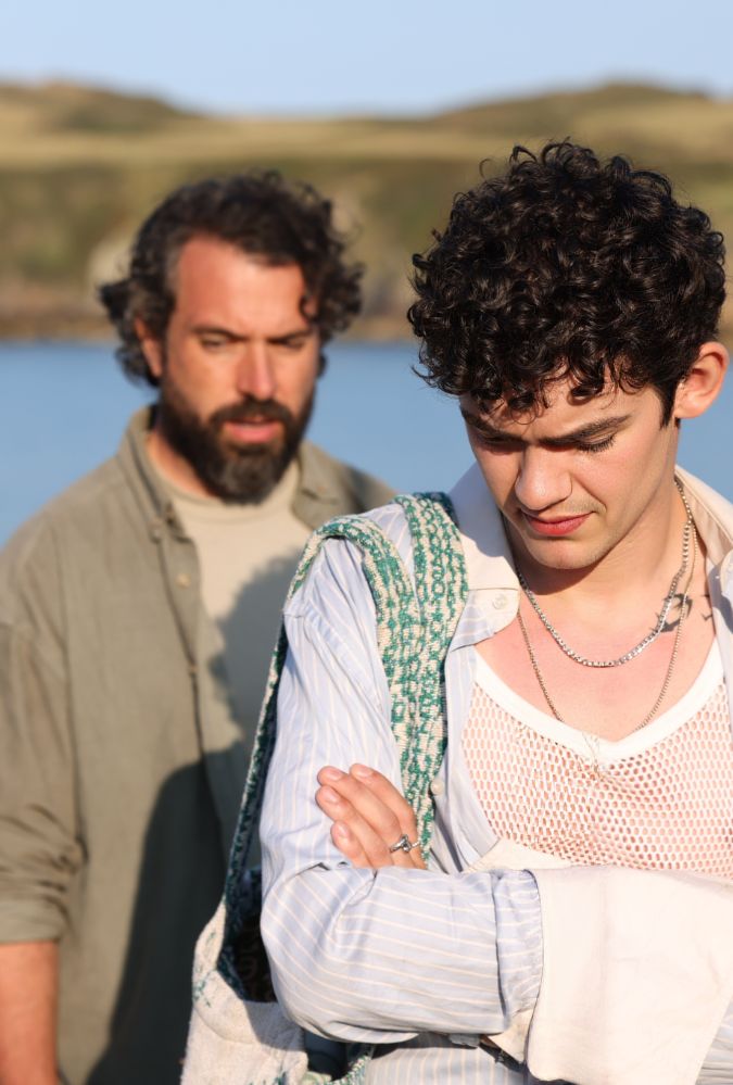A young man with curly hair stands in the foreground, arms crossed, wearing a netted shirt, striped button-down, and a necklace. In the background, an older person with a beard stands slightly blurred, looking concerned. They are outdoors near a body of water, with grassy hills in the distance under a clear sky.