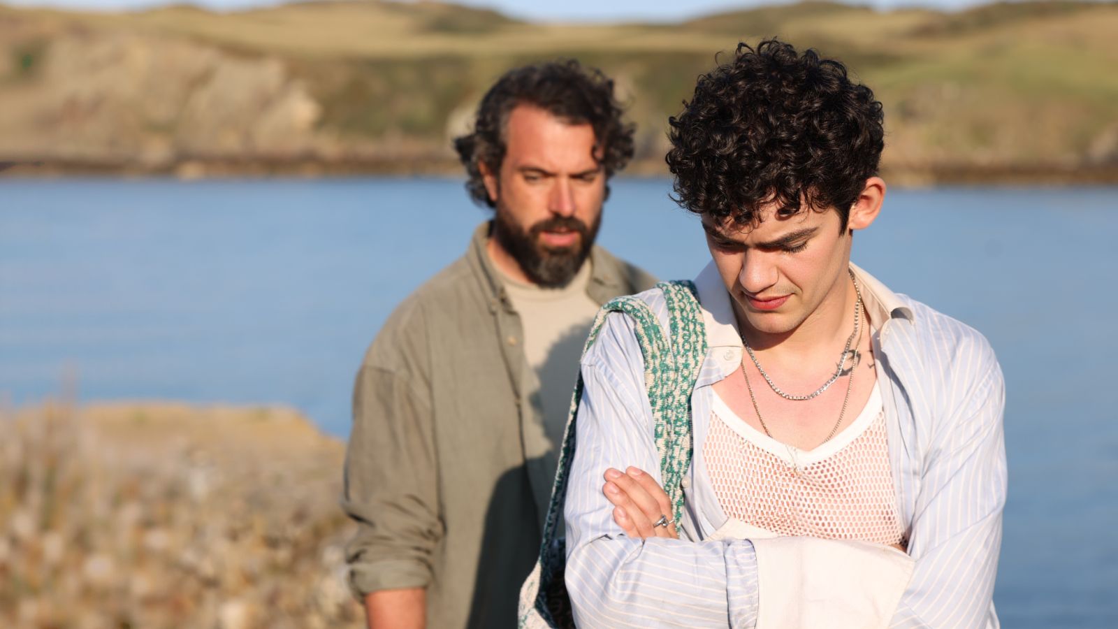 A young man with curly hair stands in the foreground, arms crossed, wearing a netted shirt, striped button-down, and a necklace. In the background, an older person with a beard stands slightly blurred, looking concerned. They are outdoors near a body of water, with grassy hills in the distance under a clear sky.