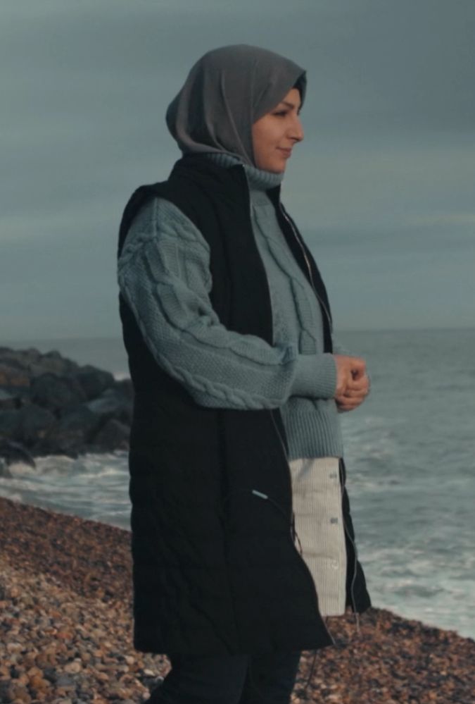 A person wearing a hijab and a dark vest over a light grey sweater stands on a pebbled beach, gazing out at the sea under an overcast sky.