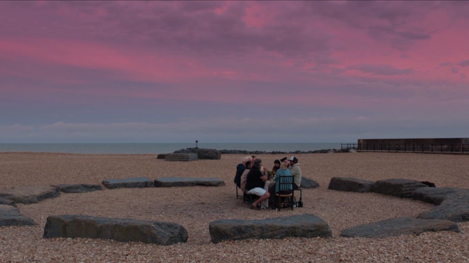 A group of people are seated at a table on a pebble beach, surrounded by large rocks, under a vibrant pink and purple sky at sunset. The ocean and horizon are visible in the background.