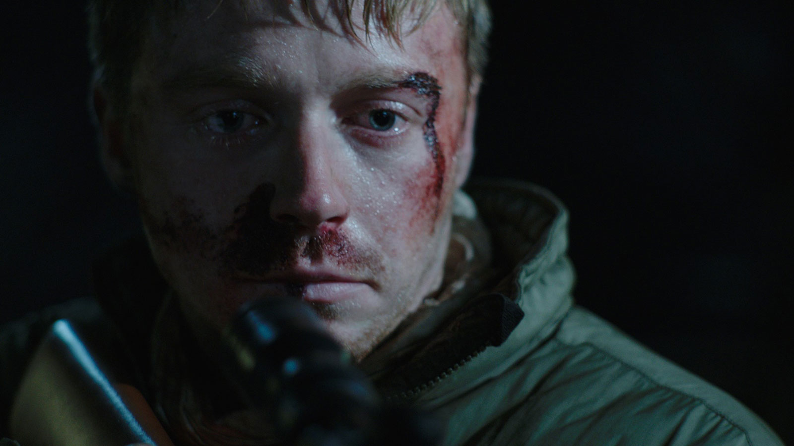 Still image from Calibre showing a man with cuts on his face holding a rifle