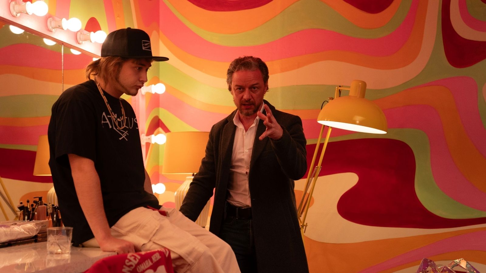Two people are in a colorful room with wavy, multicolored wall patterns. One person in a black hat and shirt sits on a vanity table, while the other, in a dark coat, gestures expressively. Brightly lit mirrors and a yellow lamp add to the vibrant atmosphere.
