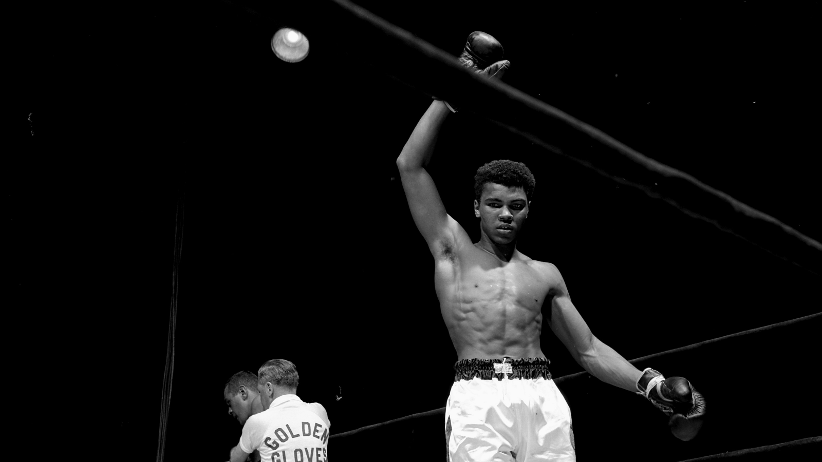 A young man, Cassius Clay, also known as Muhammad Ali, holds his right arm up in the air in victory after a boxing match. He is standing in a boxing ring, wearing white and black boxing shorts and boxing gloves.