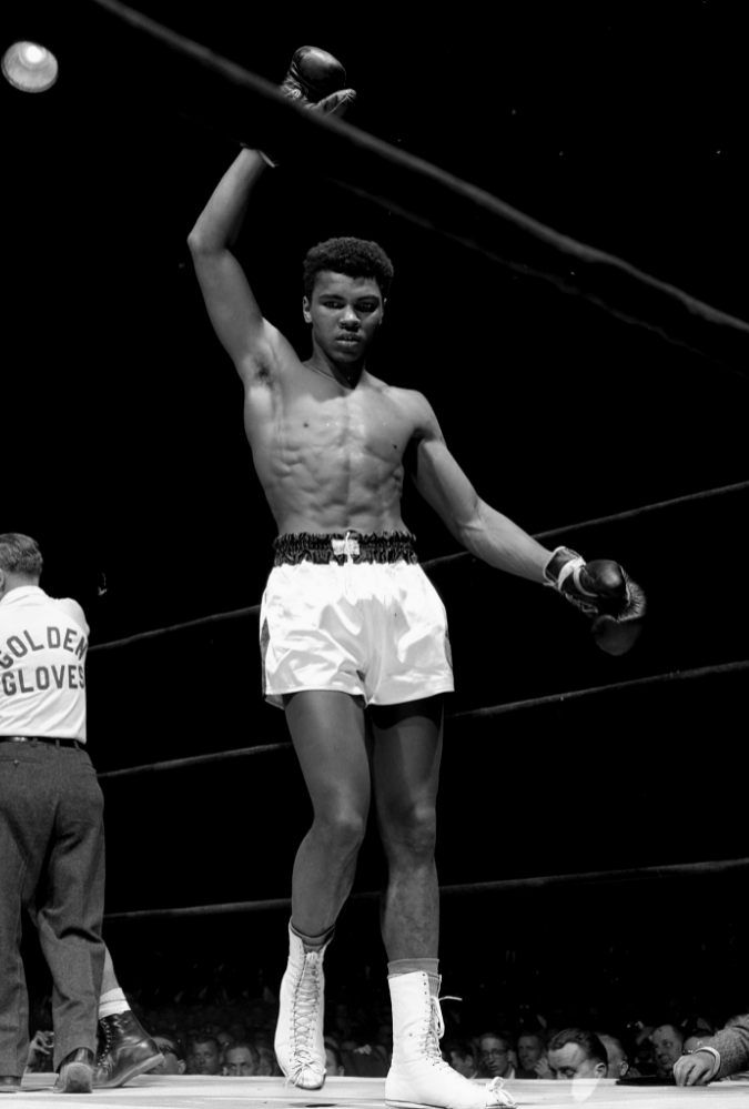 A young man, Cassius Clay, also known as Muhammad Ali, holds his right arm up in the air in victory after a boxing match. He is standing in a boxing ring surrounded by a large crowd, and is wearing white and black boxing shorts, white lace up boots and boxing gloves.