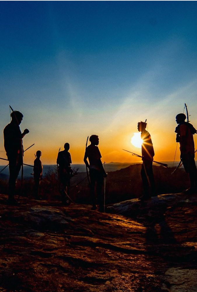 Silhouettes of seven people with bows and arrows stand on a hill against a striking sunset backdrop, with orange and blue hues blending in the sky.