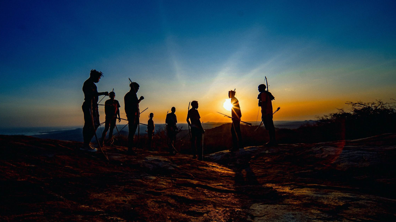 Silhouettes of seven people with bows and arrows stand on a hill against a striking sunset backdrop, with orange and blue hues blending in the sky.