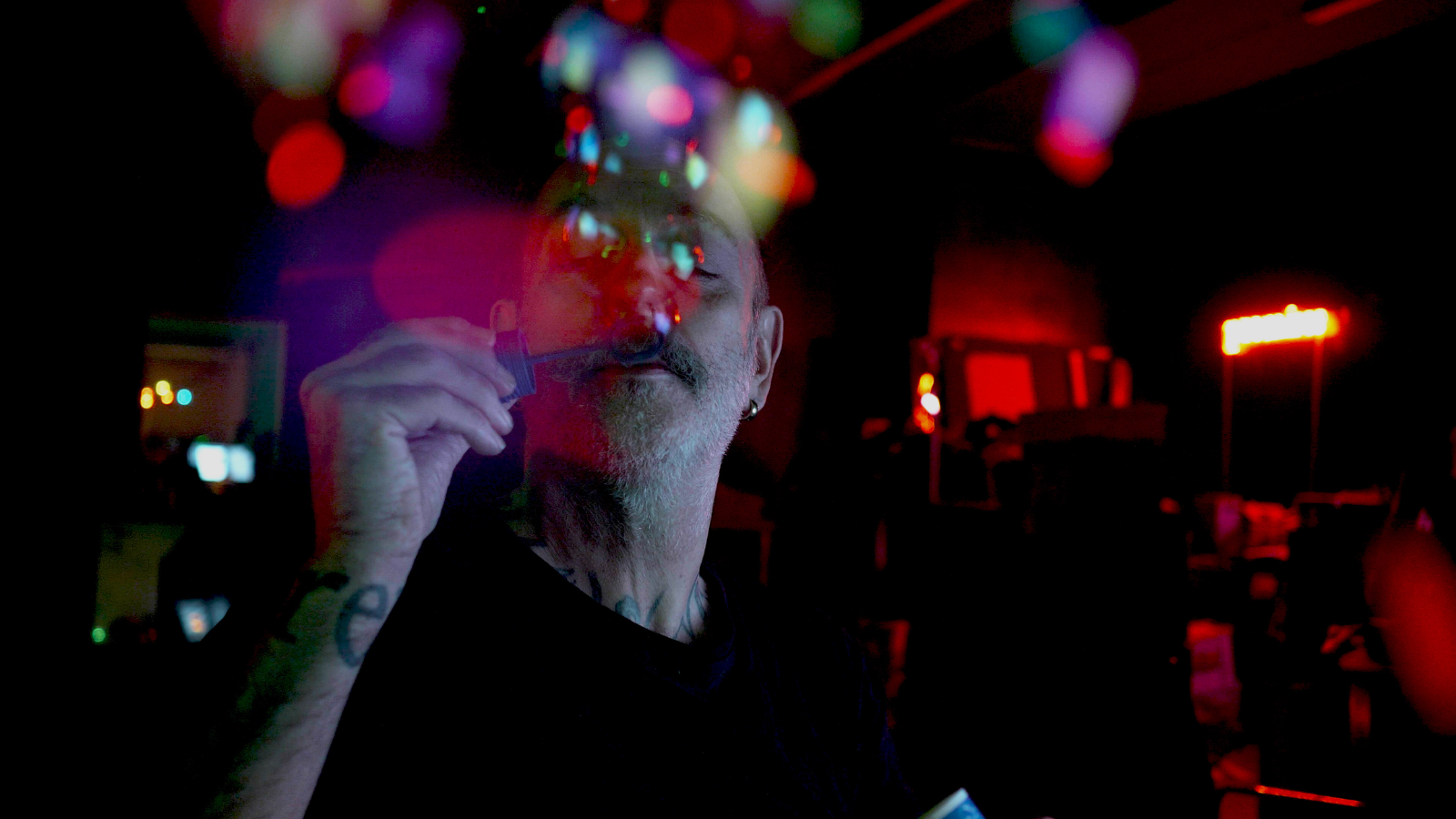 A man with a beard is blowing bubbles indoors, with colorful lights reflecting off the bubbles. The room is dark, illuminated by vibrant red, blue, and green lights, creating a visually striking effect. He holds a bubble wand close to his face and there is a laptop in front of him.