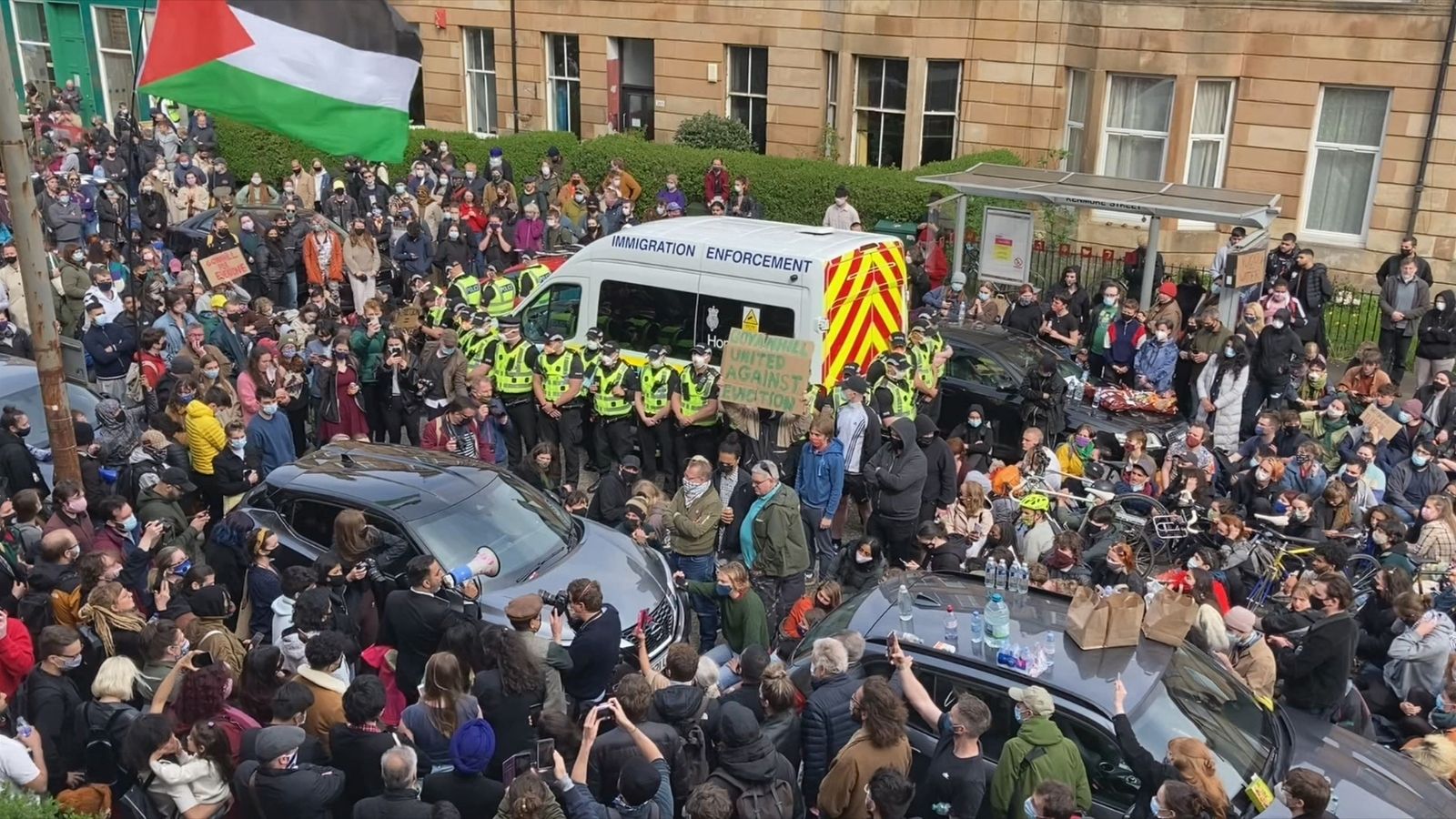 Crowd gathered around an Immigration Enforcement van, with many people standing and sitting on the street, some holding signs. A large Palestinian flag is visible. Police officers in high-visibility jackets form a line near the van. The setting appears to be urban, with a building in the background.