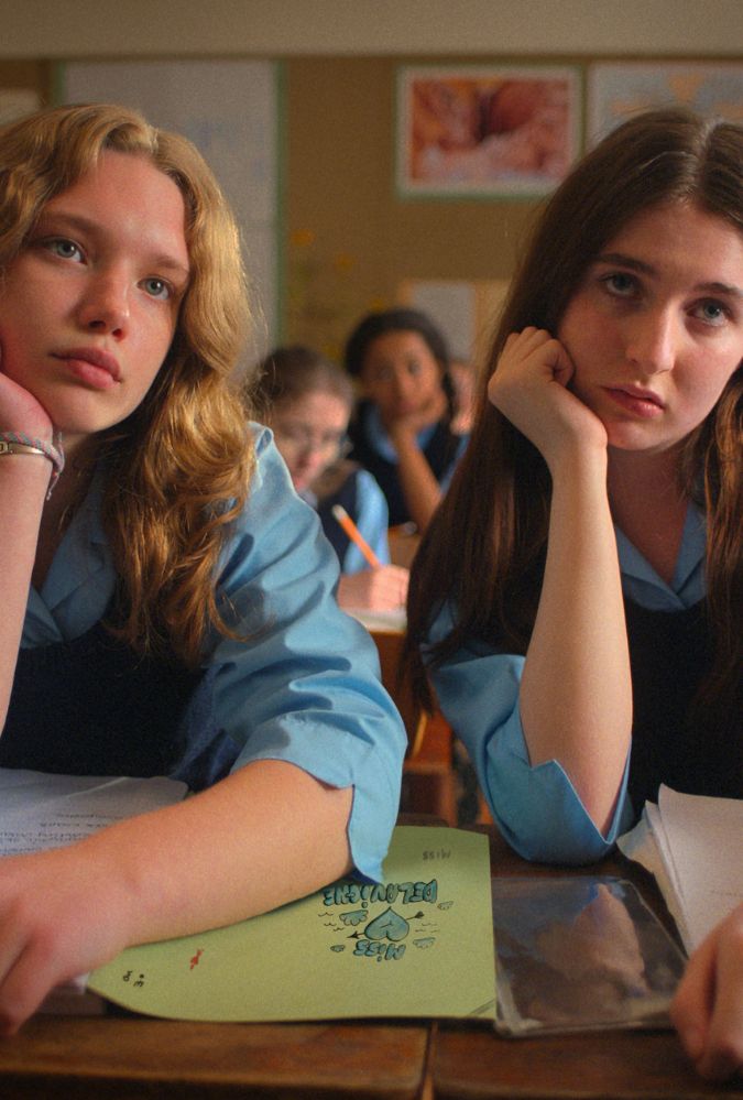 Two students in blue uniforms sit at a classroom desk, appearing bored and resting their heads on their hands. Papers and a fuzzy pink pencil case are on the desk. Other students in similar attire are in the background.