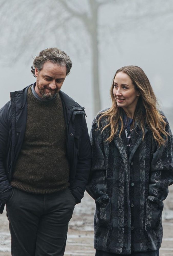 A man and a woman walk side by side on a foggy path near a body of water. They wear winter coats, and the man has his hands in his pockets, while the woman looks at him, smiling. A goose is visible on the path behind them, and blurred figures and benches are in the misty background.