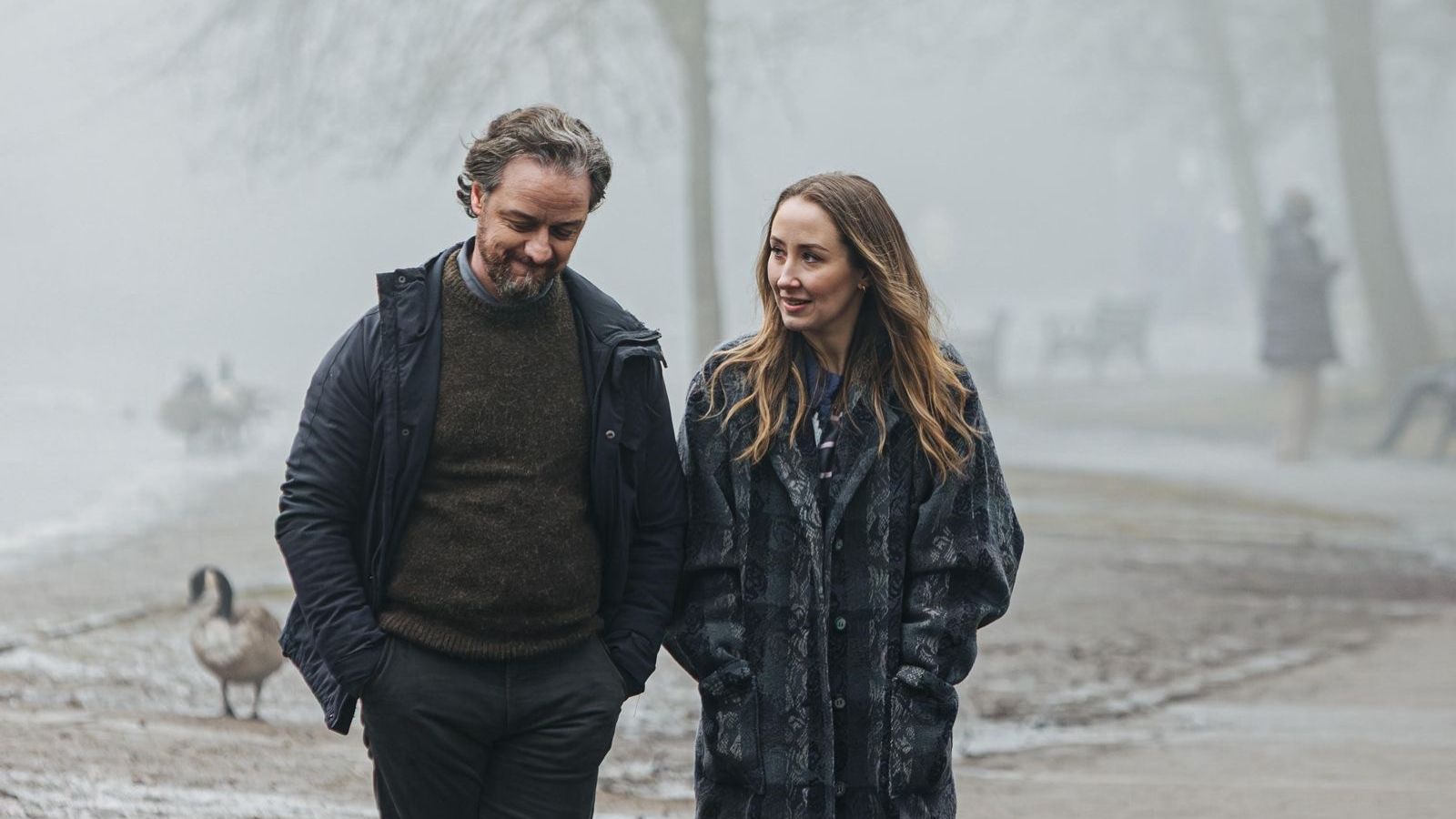 A man and a woman walk side by side on a foggy path near a body of water. They wear winter coats, and the man has his hands in his pockets, while the woman looks at him, smiling. A goose is visible on the path behind them, and blurred figures and benches are in the misty background.