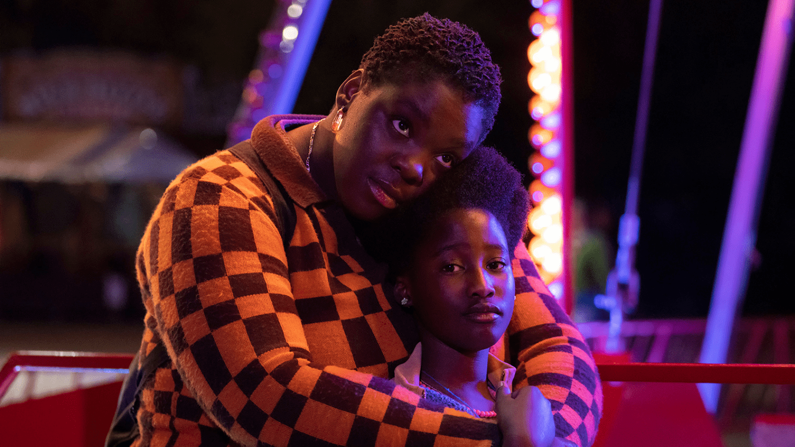 Black woman and child hug each other on fair ground ride at night lit up in neon lights