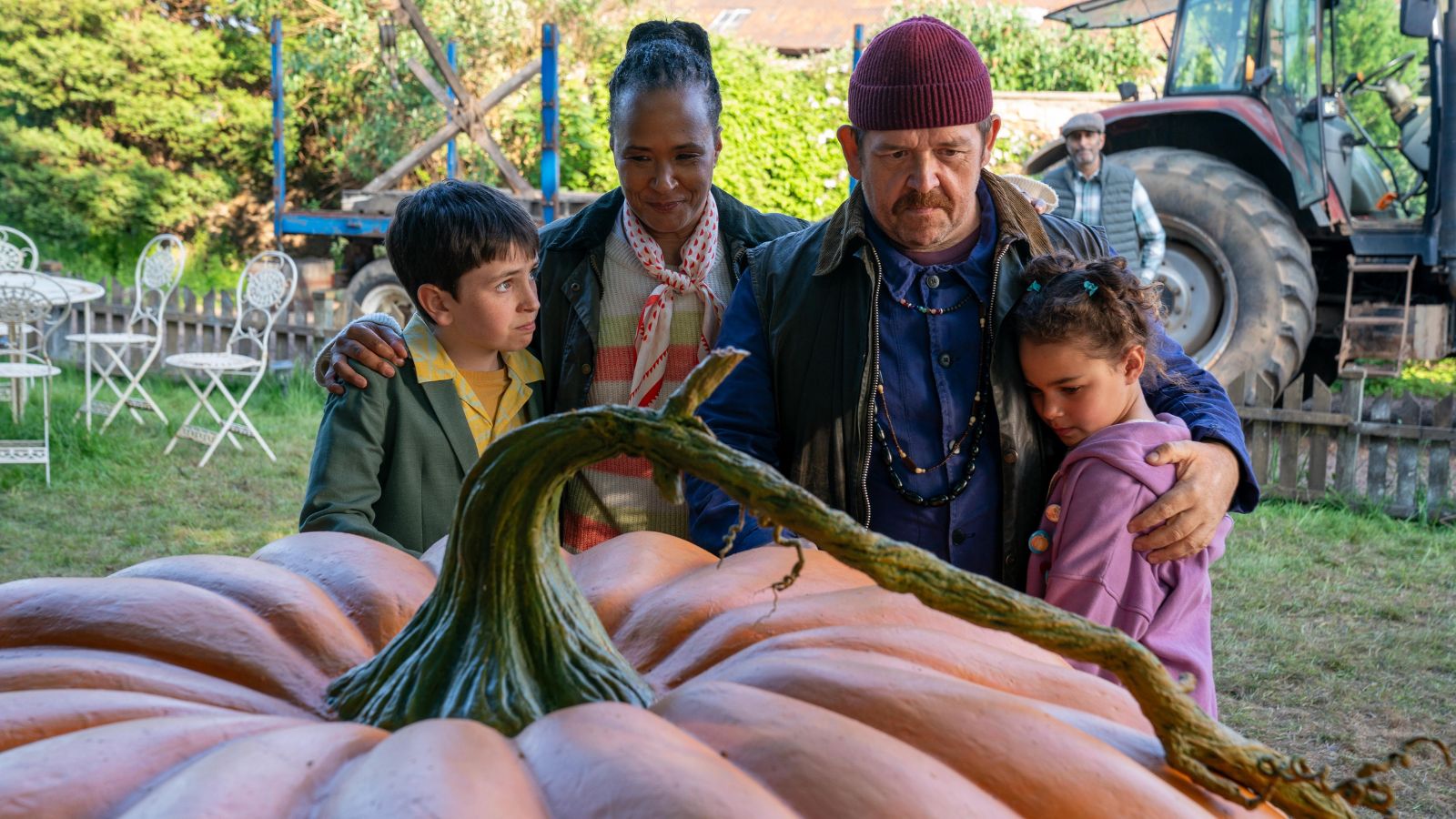 A group of four people, including a woman, a man, and two children, stand closely together around a giant pumpkin. The setting is outdoors with green foliage, a tractor, and white garden furniture in the background. The mood is warm and reflective, with the adults embracing the children near the large vegetable.