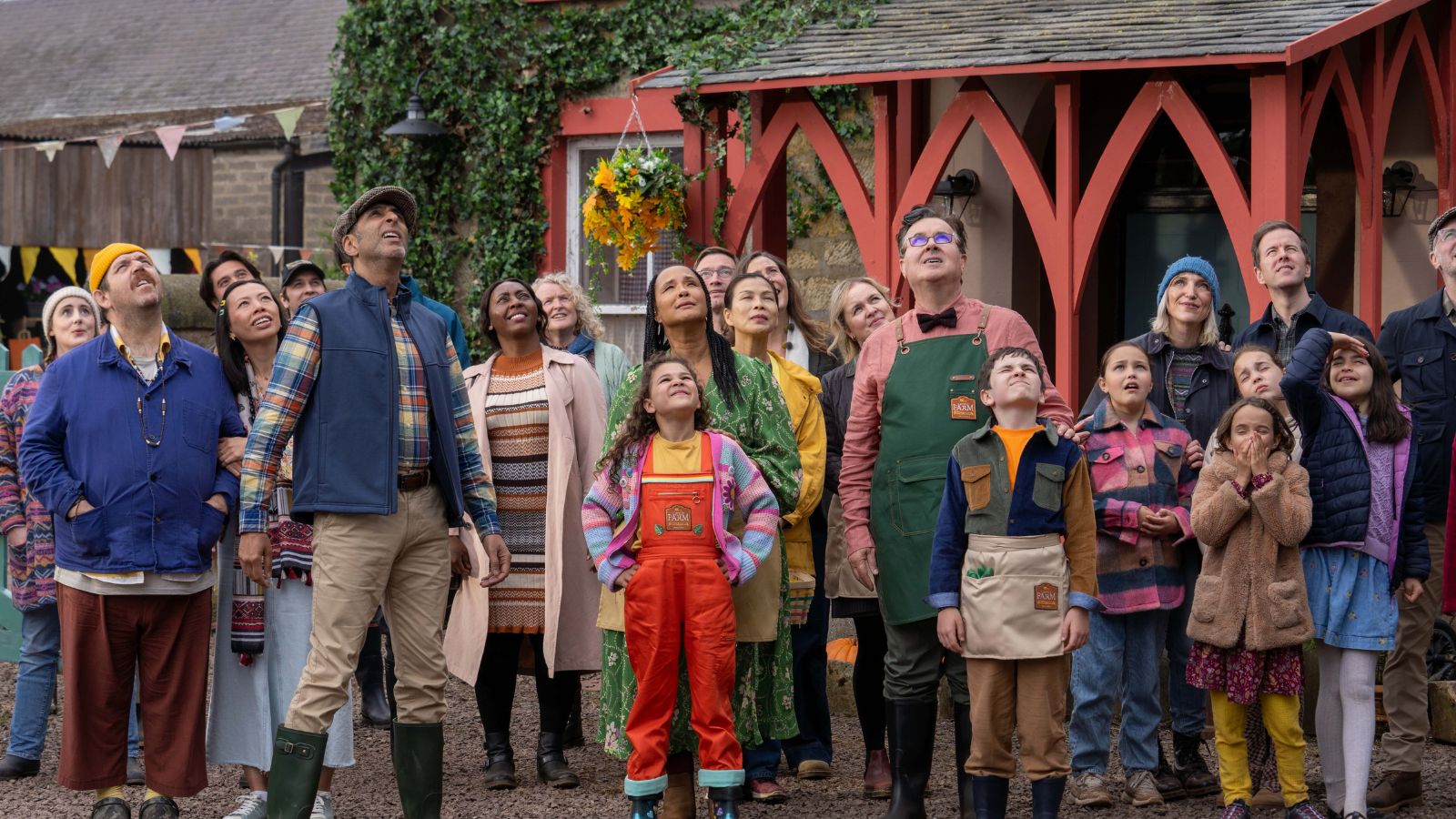 A diverse group of adults and children, dressed in colourful outfits and aprons, look upward with expressions of wonder and anticipation. They stand in front of a rustic building adorned with ivy and bunting, conveying a lively, communal atmosphere.