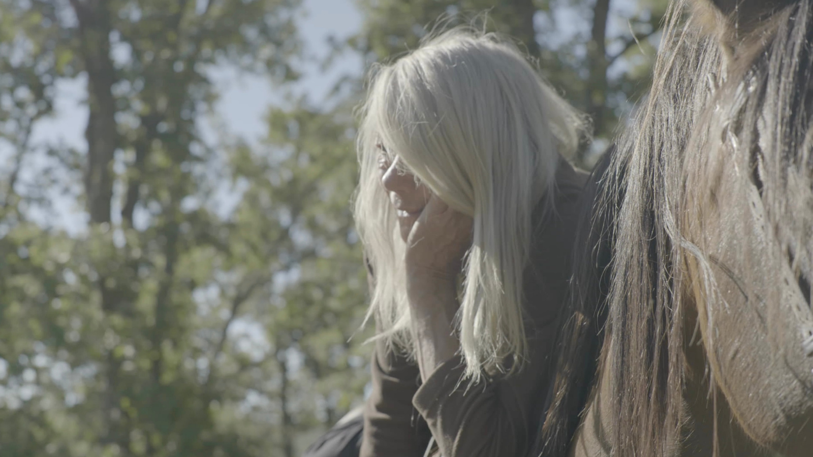 A woman with long, light hair leans against a horse, surrounded by a green, sunlit forest.